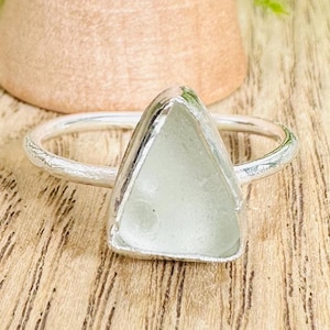Puede incluir: Un anillo de plata con una pieza triangular de vidrio de mar engastada en el centro. El anillo está sobre una superficie de madera.