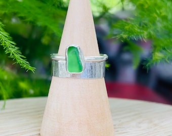 Puede incluir: Un anillo de plata con una piedra de vidrio de mar verde engastada en el centro. El anillo está en un expositor de madera.