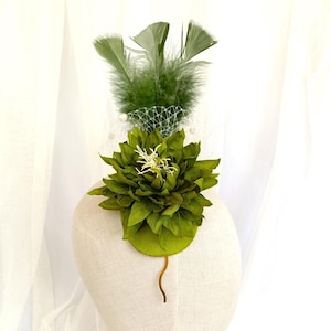 Peut inclure: Une pince à cheveux en forme de fleur verte avec une tige en fil d'or et des plumes vertes. La pince est sur une tête de mannequin blanche.