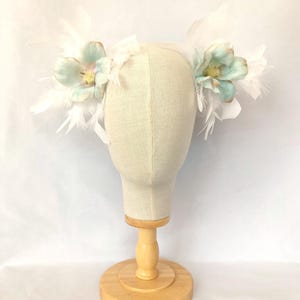 Könnte beinhalten: Ein weißes Stirnband mit zwei hellblauen Blumen und weißen Federn. Das Stirnband befindet sich auf einem Mannequin-Kopf.