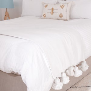 May include: White bed with a white throw blanket with pom pom trim. The throw blanket is folded at the foot of the bed.