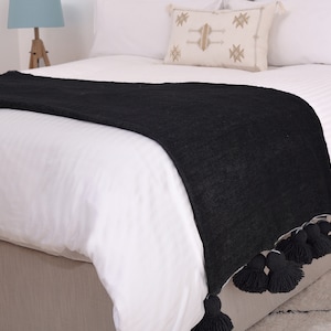 May include: Black throw blanket with a fringe of black pom-poms. The blanket is draped over a white bed with a white pillow with a geometric pattern.