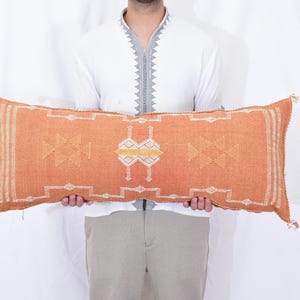 Könnte beinhalten: Ein orangefarbenes rechteckiges Kissen mit geometrischen Mustern in Weiß und Gelb. Das Kissen hat an jedem Ende Quasten. Das Kissen wird von einer Person gehalten, die ein weißes Hemd und eine khakifarbene Hose trägt.
