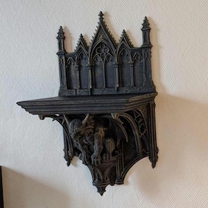 Peut inclure: Une étagère murale de style gothique, de couleur gris foncé, avec des détails architecturaux complexes. L'étagère est ornée d'une sculpture de gargouille en guise de support, ajoutant une touche fantastique. Montée sur un mur blanc texturé.