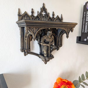 Puede incluir: Un estante de pared con acabado en color bronce oscuro y detalles arquitectónicos góticos intrincados, incluyendo arcos apuntados y agujas. Una figura esculpida de un caballero con armadura completa se arrodilla sosteniendo una espada.