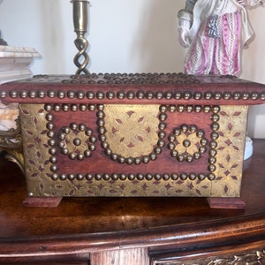 Op de afbeelding: Een houten doos met messing accenten en decoratief metaalwerk. De doos heeft een donkerhouten afwerking en is versierd met messing studs en metalen platen. De metalen platen hebben een bloemmotief.