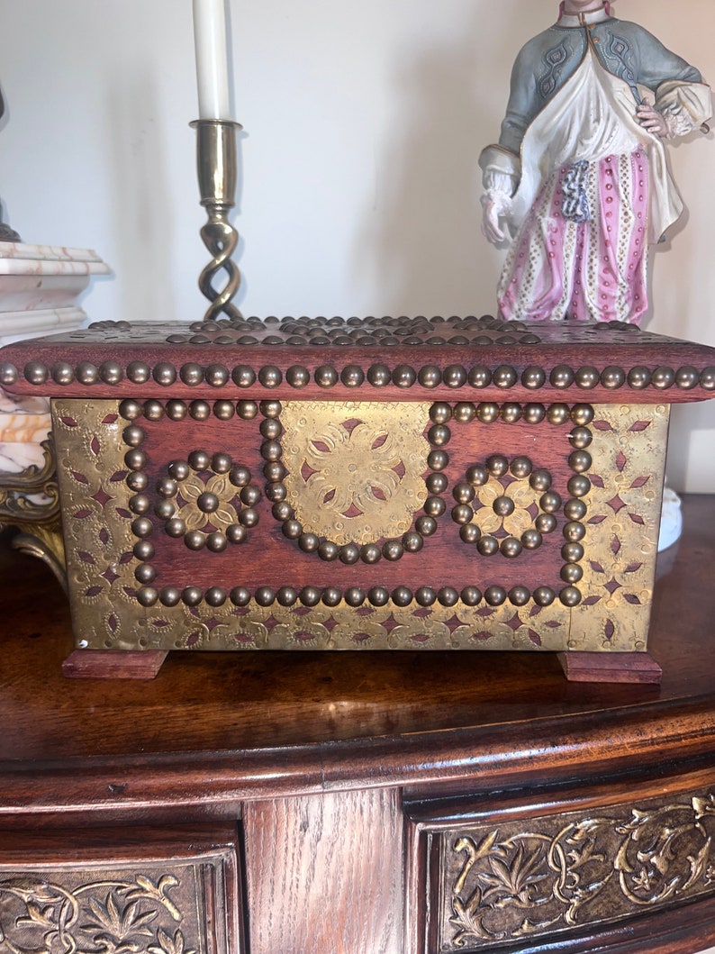 Op de afbeelding: Een houten doos met messing accenten en decoratief metaalwerk. De doos heeft een donkerhouten afwerking en is versierd met messing studs en metalen platen. De metalen platen hebben een bloemmotief.
