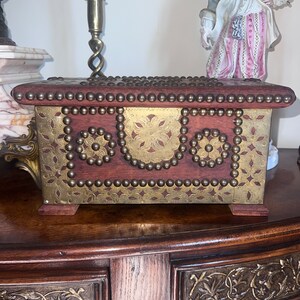 Op de afbeelding: Een houten kist met messing accenten en decoratieve studs. De kist heeft een scharnierend deksel en is versierd met een bloemmotief. De kist staat op een houten tafel.