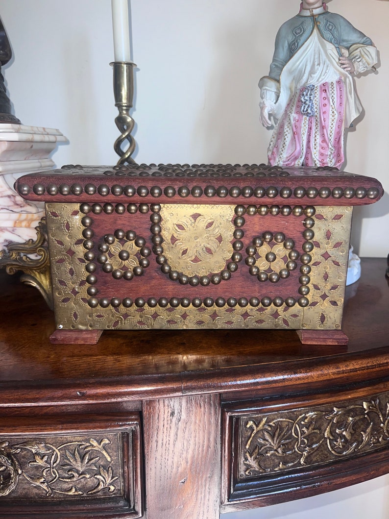 Op de afbeelding: Een houten kist met messing accenten en decoratieve studs. De kist heeft een scharnierend deksel en is versierd met een bloemmotief. De kist staat op een houten tafel.