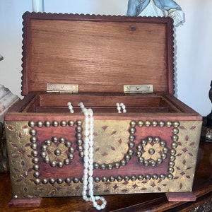 Op de afbeelding: Een houten doos met messing accenten en een parelketting erin. De doos heeft een decoratief ontwerp met bloemmotieven en is versierd met messing studs. De doos is open en de ketting hangt erin.