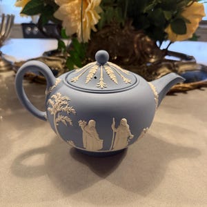 Może przedstawiać: Jasnoniebieski ceramiczny imbryk z pokrywką i uchwytem. Imbryk posiada białe reliefowe wzory postaci i drzew. Imbryk stoi na jasnoszarej powierzchni, z kompozycją kwiatową w tle.