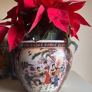 May include: A red poinsettia plant in a white ceramic pot with a blue and gold rim. The pot is decorated with a painted scene of people in traditional clothing and a horse.