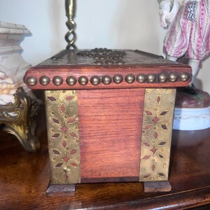 Op de afbeelding: Een houten doos met messing accenten en decoratief metaalwerk. De doos heeft een donkerhouten afwerking en is versierd met messing studs en een messing rand rond de bovenrand. De doos heeft een rechthoekige vorm en staat op een houten oppervlak.