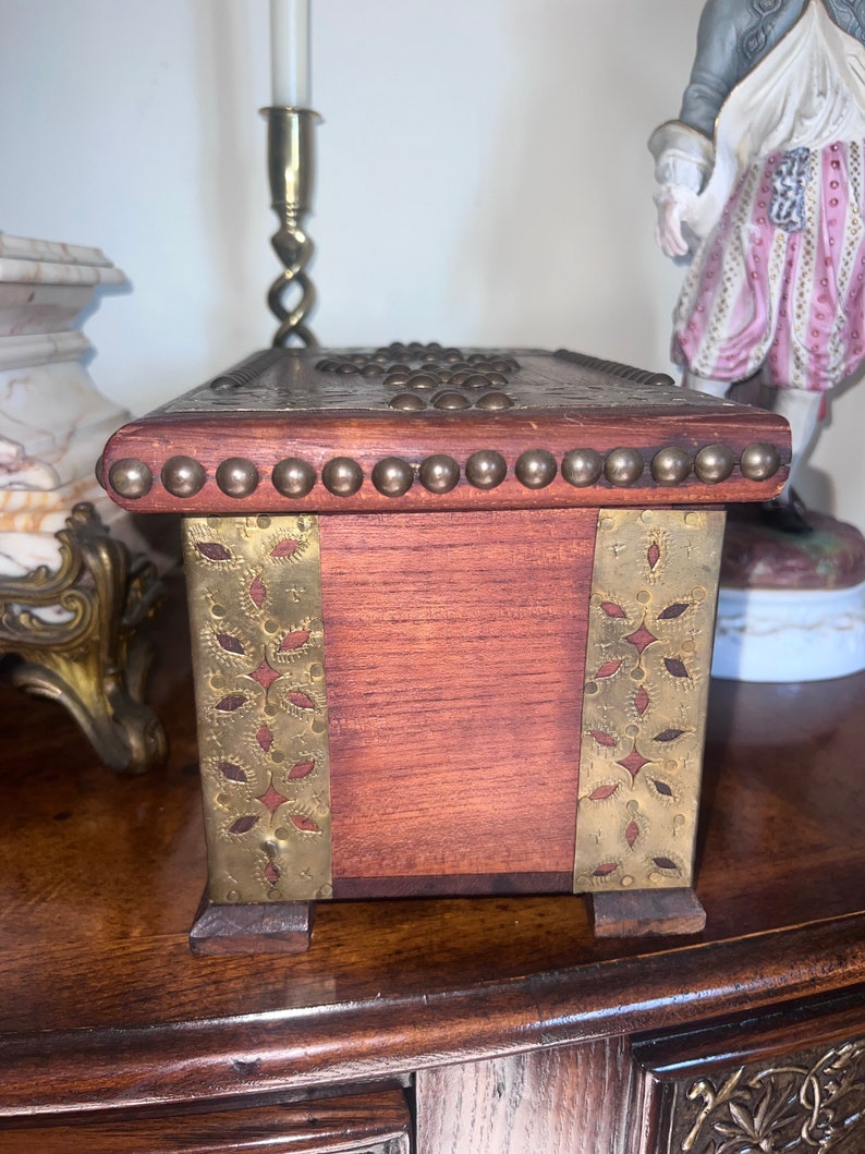 Op de afbeelding: Een houten doos met messing accenten en decoratief metaalwerk. De doos heeft een donkerhouten afwerking en is versierd met messing studs en een messing rand rond de bovenrand. De doos heeft een rechthoekige vorm en staat op een houten oppervlak.