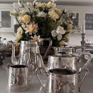 May include: A silver tea set consisting of two teapots, a creamer, and a sugar bowl. Each piece features a polished metallic finish, an engraved leaf wreath design on the body, and a decorative scalloped border near the top rim.