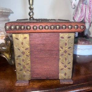 Op de afbeelding: Een kleine houten doos met messing accenten. De doos heeft een decoratief ontwerp aan de voor- en zijkanten. De doos is gemaakt van donker hout en heeft een messing rand rond de bovenkant.