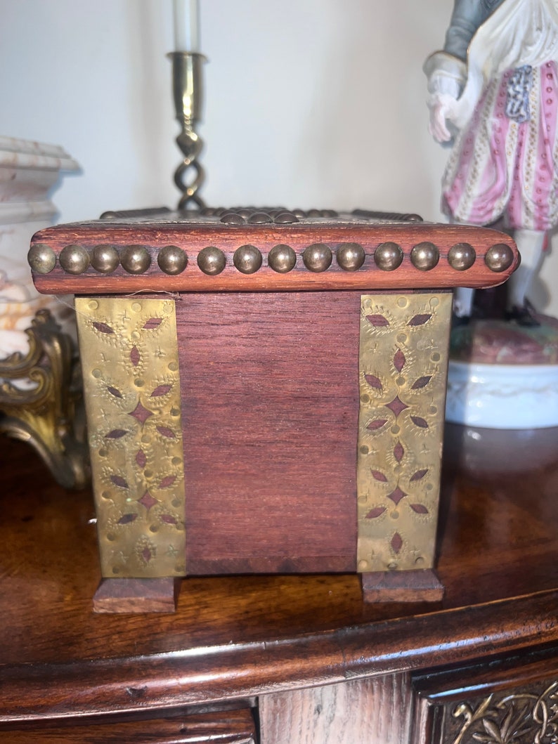 Op de afbeelding: Een kleine houten doos met messing accenten. De doos heeft een decoratief ontwerp aan de voor- en zijkanten. De doos is gemaakt van donker hout en heeft een messing rand rond de bovenkant.