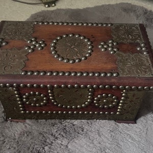 Op de afbeelding: Een houten doos met ingewikkelde metalen ontwerpen en studs. De doos is donkerbruin met gouden accenten en heeft een scharnierend deksel.