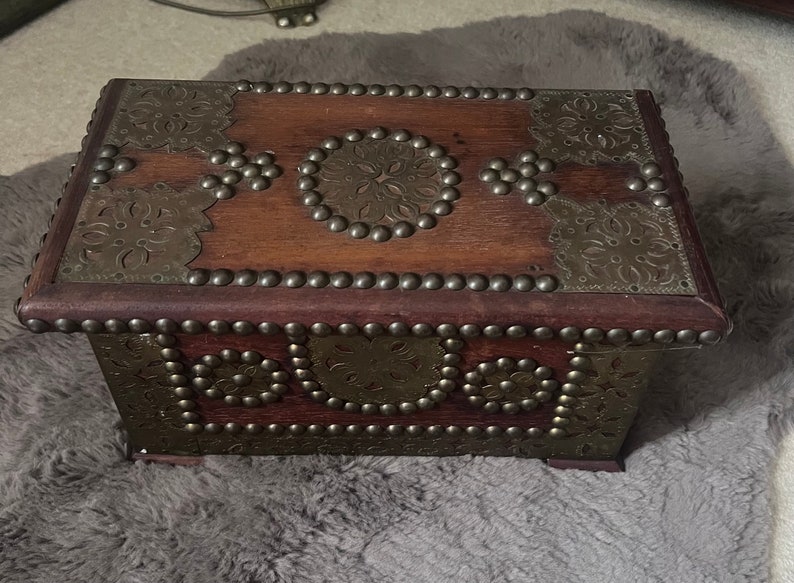 Op de afbeelding: Een houten doos met ingewikkelde metalen ontwerpen en studs. De doos is donkerbruin met gouden accenten en heeft een scharnierend deksel.