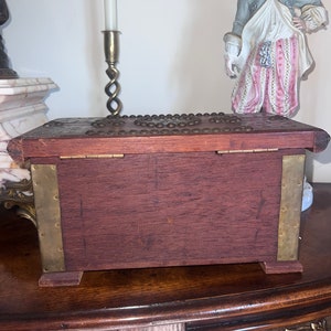 Op de afbeelding: Een houten kist met messing accenten en een decoratief deksel. De kist is gesloten en staat op een houten tafel.