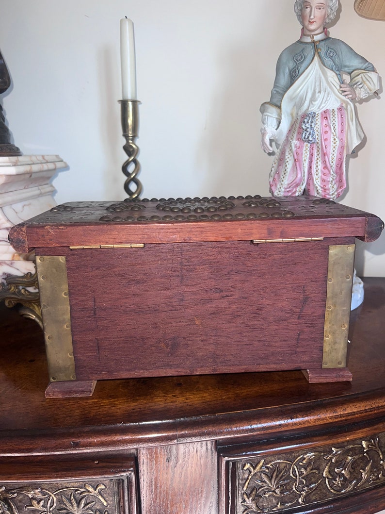 Op de afbeelding: Een houten kist met messing accenten en een decoratief deksel. De kist is gesloten en staat op een houten tafel.
