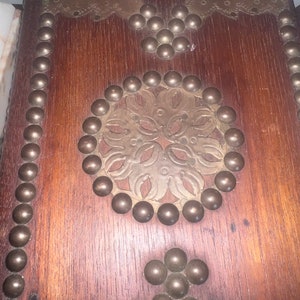 Op de afbeelding: Een houten kist met een decoratief messing ontwerp en metalen studs. De kist heeft een donkerbruine afwerking en is versierd met een bloemmotief.