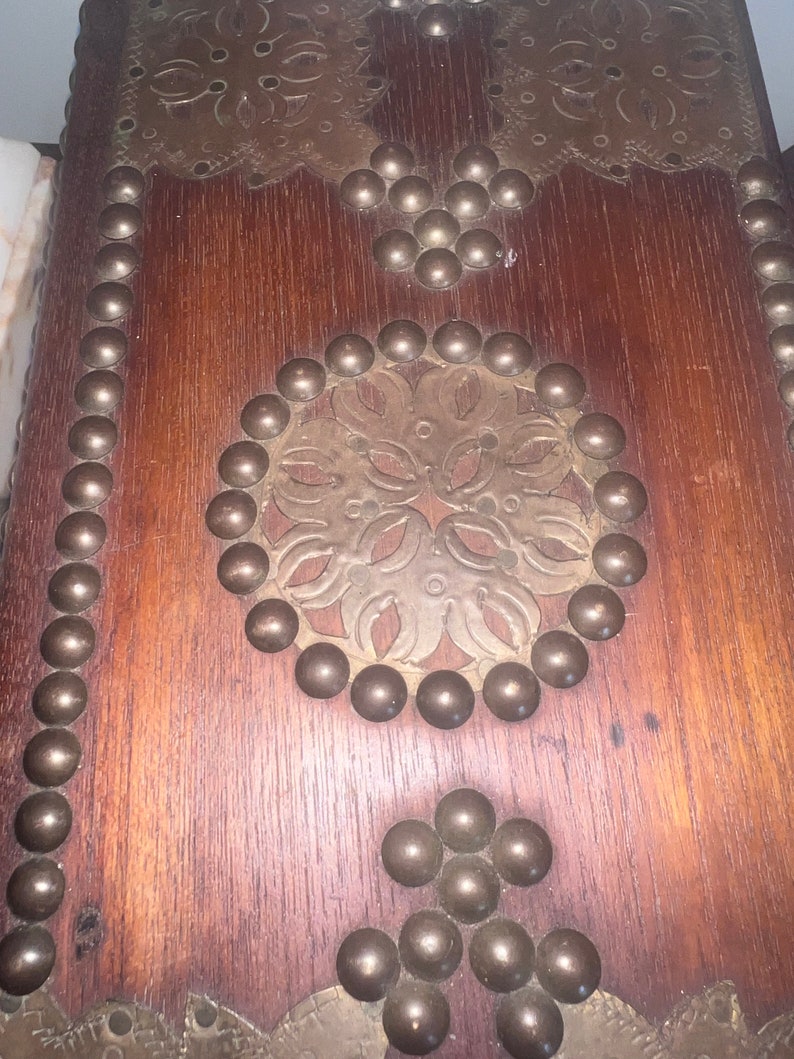 Op de afbeelding: Een houten kist met een decoratief messing ontwerp en metalen studs. De kist heeft een donkerbruine afwerking en is versierd met een bloemmotief.
