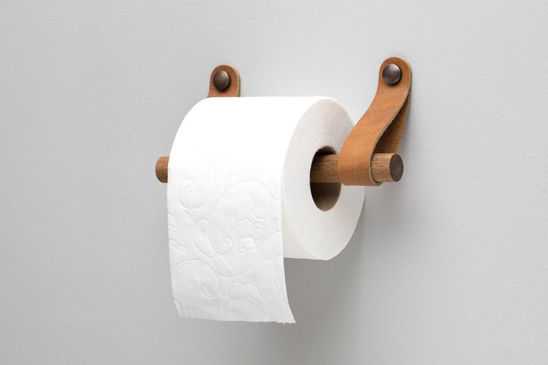 May include: A wooden toilet paper holder with brown leather straps and metal rivets. The holder is mounted to a white wall and has a roll of white toilet paper on it.