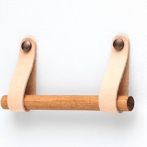 May include: A wooden towel bar with leather straps and brass rivets. The bar is mounted to a white wall.