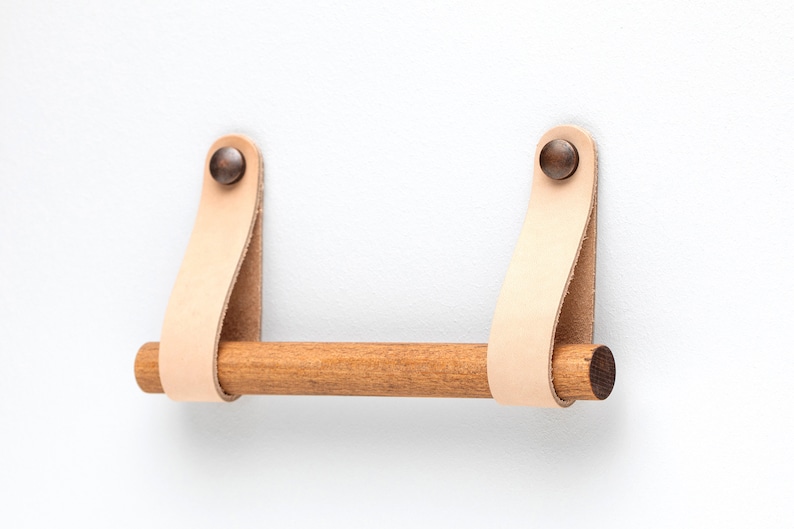 May include: A wooden towel bar with leather straps and brass rivets. The bar is mounted to a white wall.
