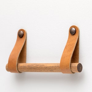 May include: A wooden towel bar with leather straps and brass accents. The bar is mounted to a white wall.