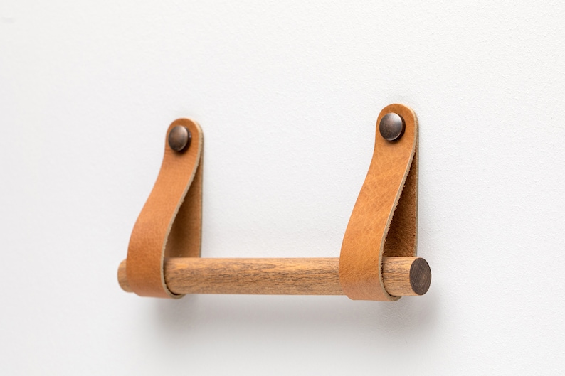 May include: A wooden towel bar with leather straps and brass accents. The bar is mounted to a white wall.