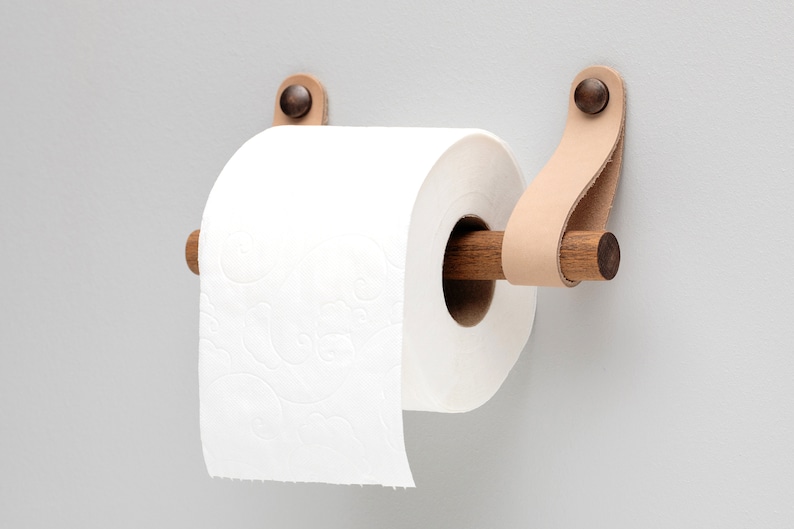 May include: A wooden toilet paper holder with leather straps and a brown metal accent. The holder is mounted to a wall and has a roll of white toilet paper on it.
