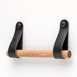 May include: A wooden towel bar with black leather straps and metal rivets. The bar is mounted to a white wall.