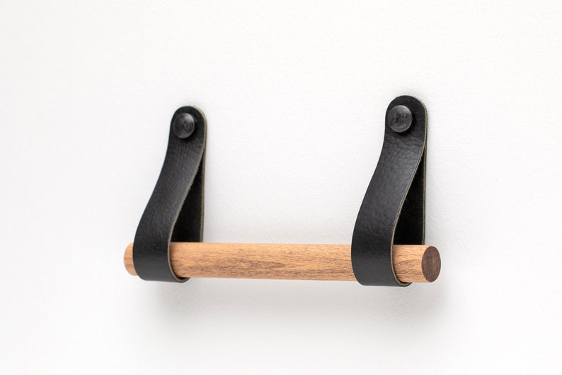 May include: A wooden towel bar with black leather straps and metal rivets. The bar is mounted to a white wall.