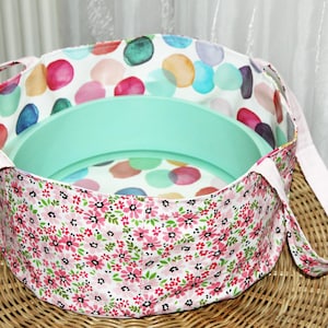 Könnte beinhalten: Ein runder Stoffkorb mit floralem Muster außen und einem bunten Tupfenmuster innen. Der Korb hat hellrosa Griffe und ein hellgrünes Tablett im Inneren.