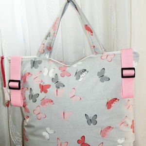 Könnte beinhalten: Eine graue Tragetasche mit einem wiederkehrenden Muster aus rosa, weißen und dunkelgrauen Schmetterlingen. Die Tasche verfügt über zwei graue Stoffgriffe, einen soliden schwarzen Boden und zwei verstellbare rosa Seitengurte mit schwarzen Kunststoffschnallen.