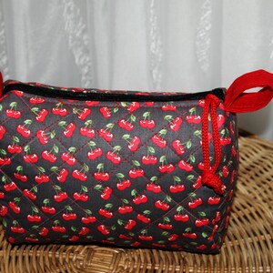 Könnte beinhalten: Eine rechteckige, gesteppte Tasche mit einem Muster aus roten Kirschen auf schwarzem Grund. Die Tasche hat rote Griffe und einen roten Kordelzugverschluss. Sie liegt auf einer Korboberfläche.