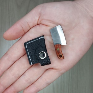 Può includere: Un piccolo fodero per coltello in pelle nera con chiusura a scatto. Il fodero è appoggiato su una mano, accanto a un piccolo coltello con manico in legno e argento.