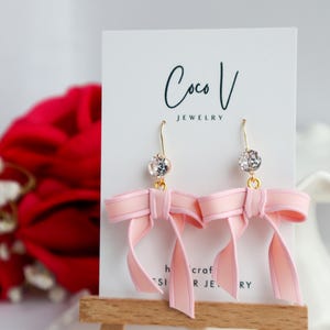May include: A pair of pink bow earrings with a gold hook and a clear crystal ball. The earrings are displayed on a white card with the text "Coco V Jewelry". A red rose is in the background.