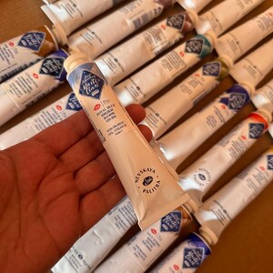 May include: A hand holding a tube of Royal Blue oil paint from the Nevskaya Palitra Master Class series. The tube is white with blue and gold accents. Other tubes of oil paint are visible in the background.
