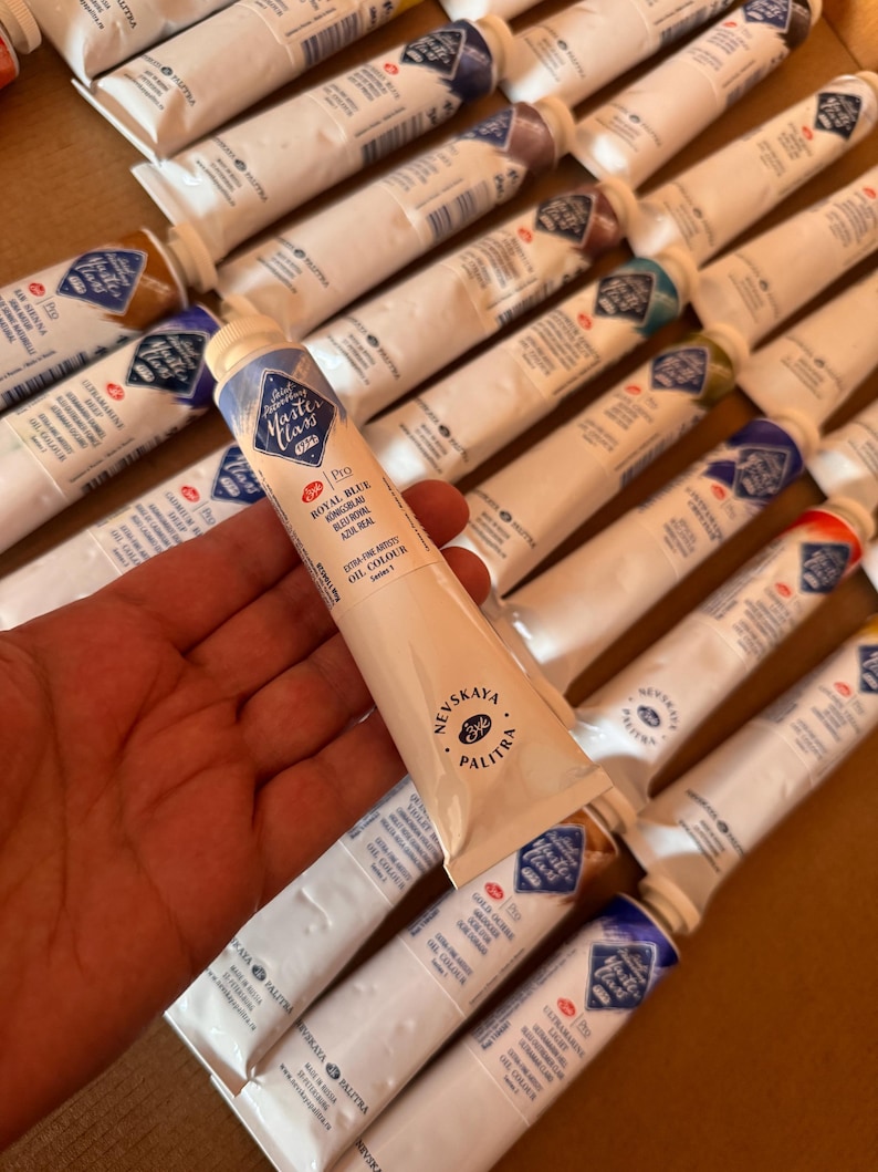 May include: A hand holding a tube of Royal Blue oil paint from the Nevskaya Palitra Master Class series. The tube is white with blue and gold accents. Other tubes of oil paint are visible in the background.
