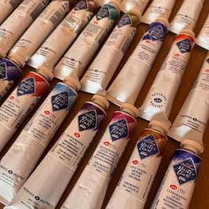 May include: An assortment of white oil paint tubes with colorful labels, arranged closely together. The labels display the brand "Master Class" and various color names like "Ceruleum Blue" and "Ultramarine Light". The tubes are on a brown surface.