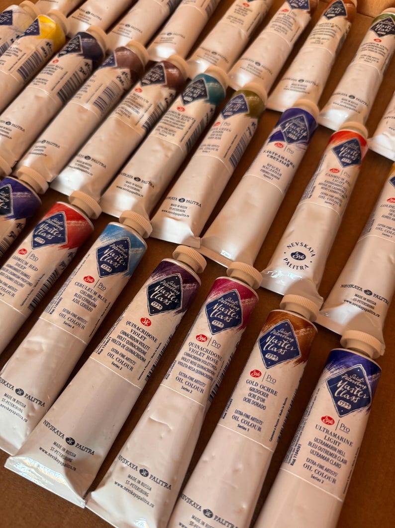 May include: An assortment of white oil paint tubes with colorful labels, arranged closely together. The labels display the brand "Master Class" and various color names like "Ceruleum Blue" and "Ultramarine Light". The tubes are on a brown surface.
