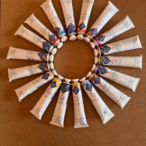 May include: A circular arrangement of white paint tubes with colorful caps, including blue, red, and yellow. The tubes are arranged on a brown surface, creating a sunburst effect. The product is likely art supplies.