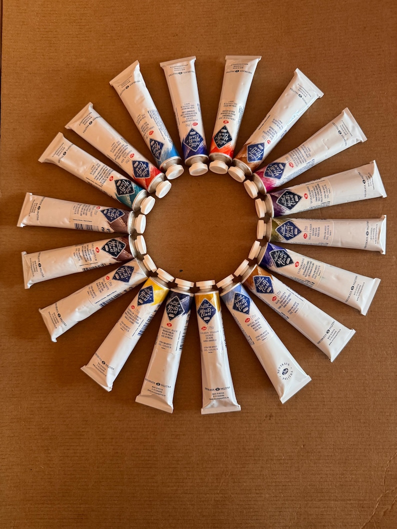 May include: A circular arrangement of white paint tubes with colorful caps, including blue, red, and yellow. The tubes are arranged on a brown surface, creating a sunburst effect. The product is likely art supplies.