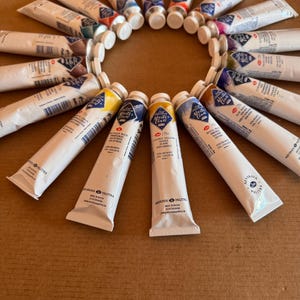 May include: A collection of white paint tubes arranged in a circular pattern. The tubes have colorful caps and labels with text. The paint tubes are labeled "Master Class" and contain various colors, including yellow, blue, and red. The background is a light brown surface.