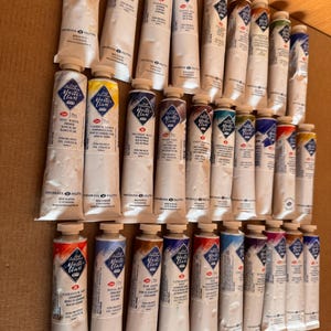 May include: A collection of oil paint tubes in various colors, including white, yellow, blue, and red. The tubes are labeled with the brand name "Master Class" and the color names. The tubes are arranged on a light brown surface.