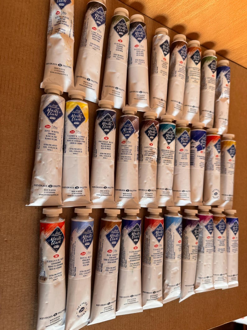 May include: A collection of oil paint tubes in various colors, including white, yellow, blue, and red. The tubes are labeled with the brand name "Master Class" and the color names. The tubes are arranged on a light brown surface.