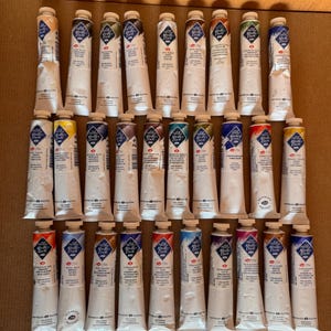 May include: A collection of artist paint tubes, each with a white body and a colored cap. The tubes are arranged in rows, showcasing a variety of colors. The labels feature text and a diamond-shaped logo. The paint tubes are on a brown surface.